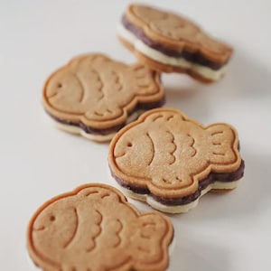 May include: Four fish-shaped cookies with a light brown exterior and a detailed fish design. The cookies have a dark filling and a white layer, creating a sandwich effect. The cookies are arranged on a white surface.