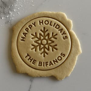 May include: A circular cookie dough cutout featuring an embossed snowflake design in the center. The text HAPPY HOLIDAYS is arched across the top, and the text THE BIFANOS is arched across the bottom, all pressed into the light beige dough.