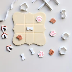 May include: A collection of dog-themed cookie cutters and a tic-tac-toe board made of dough. The cutters include paw prints, dog bones, dog faces, and house shapes. The tic-tac-toe board has dog-themed pieces in place.