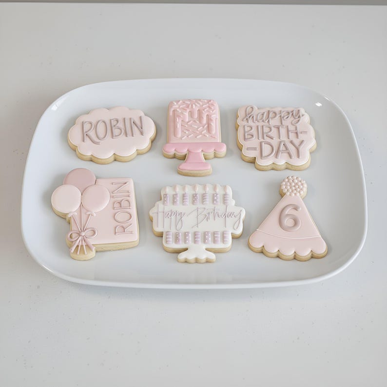 Pode incluir: Uma travessa branca com seis biscoitos de a&ccedil;&uacute;car decorados. Os biscoitos t&ecirc;m a forma de uma nuvem, bolo, chap&eacute;u de festa e ret&acirc;ngulos, com as palavras "Robin" e "Happy Birthday" em cinza. Os biscoitos s&atilde;o decorados em rosa e branco.