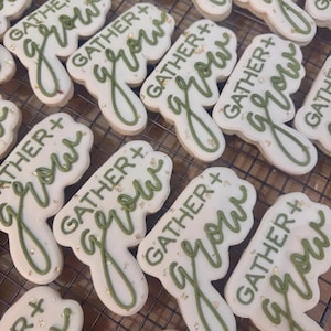 Puede incluir: Vista a&eacute;rea de varias galletas blancas con glaseado verde. Las galletas tienen forma de hoja y llevan la inscripci&oacute;n "GATHER + grow" en verde. Copos dorados espolvoreados sobre las galletas.