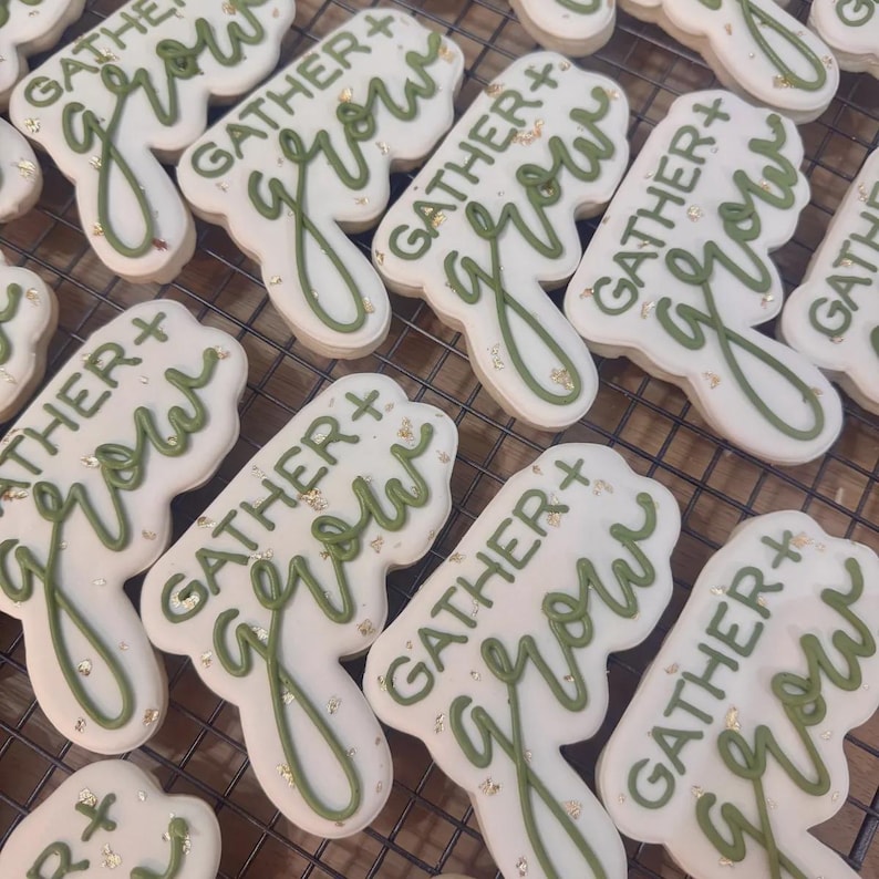 Puede incluir: Vista a&eacute;rea de varias galletas blancas con glaseado verde. Las galletas tienen forma de hoja y llevan la inscripci&oacute;n "GATHER + grow" en verde. Copos dorados espolvoreados sobre las galletas.