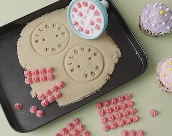 May include: A baking scene with a cookie stamp that says "I LOVE YOU" on a tray with cookie dough. Pink letter stamps and cupcakes are also present. The stamp has a light blue handle and a white stamp head.