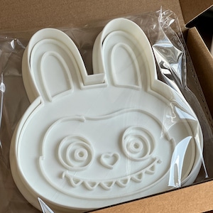 Puede incluir: Cortador de galletas blanco con forma de conejito con una cara de dibujos animados, que incluye ojos en espiral, una nariz en forma de coraz&oacute;n y una boca dentada. El cortador est&aacute; empaquetado en una bolsa de pl&aacute;stico transparente dentro de una caja de cart&oacute;n.