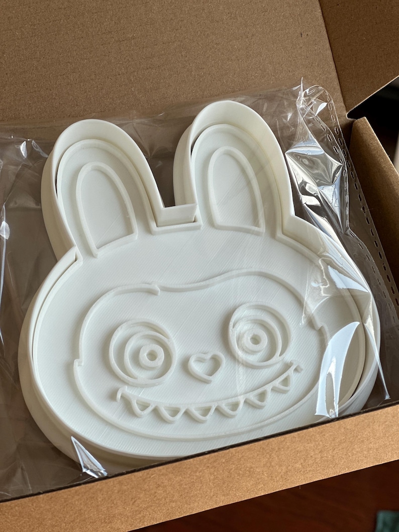 Puede incluir: Cortador de galletas blanco con forma de conejito con una cara de dibujos animados, que incluye ojos en espiral, una nariz en forma de coraz&oacute;n y una boca dentada. El cortador est&aacute; empaquetado en una bolsa de pl&aacute;stico transparente dentro de una caja de cart&oacute;n.