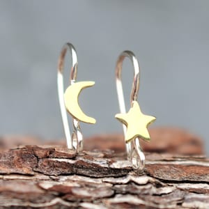 May include: A pair of silver-toned earrings with gold-colored celestial charms. One earring features a crescent moon, and the other has a five-pointed star. The earrings are displayed on a piece of wood.