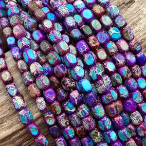 May include: Close-up of strands of gemstone beads. The beads are a mix of purple, teal, and brown, with a mottled pattern. The beads are rectangular and appear to be strung together, ready for jewelry making.
