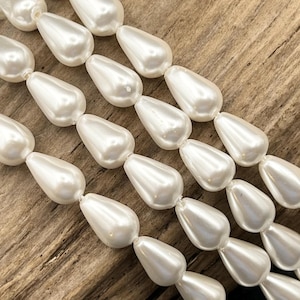 May include: A strand of white, teardrop-shaped beads with a smooth, lustrous finish. The beads are arranged in multiple parallel rows, each featuring a uniform pear-like shape and a reflective surface that catches the light, creating a consistent appearance.
