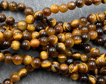 Pode incluir: Close-up de fios de contas de pedra olho de tigre. As contas redondas, castanhas e amarelo dourado, apresentam um efeito chatoyant. São enfiadas, perfeitas para a criação de joias. Cada conta tem cerca de 0,8 cm de diâmetro.