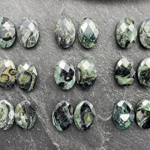 May include: Eighteen oval-shaped, faceted gemstones arranged in nine pairs. Each stone features a marbled pattern of deep forest green, black, and olive tones with a polished, reflective surface that catches the light across its geometric facets.