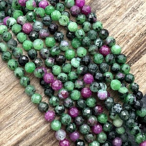 May include: Strands of faceted gemstone beads in shades of green, deep purple, and black. The beads are strung together, creating a textured and colorful display. Ideal for jewelry making.