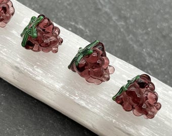 May include: Four small, translucent, dark red glass grape clusters with green leaves. The grapes are arranged on a white, rectangular surface, with a dark gray background. The grapes are detailed and appear to be hand-crafted.