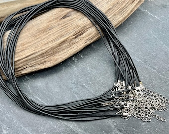 May include: A collection of black cord necklaces with silver-colored clasps and chain extenders. The cords are neatly arranged against a backdrop of weathered wood and a dark gray surface, suitable for jewelry making.