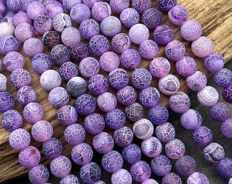 Può includere: Una collezione di perline rotonde in agata crepata viola disposte in più fili paralleli. Ogni perlina presenta un distinto motivo a ragnatela di sottili linee bianche sulla superficie, creando un aspetto strutturato in tutto il set.