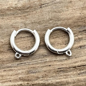 May include: Two silver-colored hoop earrings with a hinged closure and a small loop at the bottom for attaching charms or other embellishments. The earrings are displayed on a wooden surface.