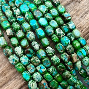 May include: Close-up of strands of green and blue gemstone beads. The beads are rectangular with rounded edges and have a mottled pattern. The color palette includes shades of green, teal, and brown. The beads are displayed on a wooden surface.