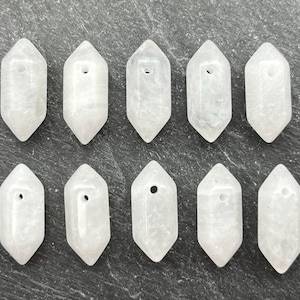 May include: Ten clear quartz crystal pendants with a hexagonal shape and a drilled hole at the top. The crystals are a translucent white color and are arranged in two rows of five on a dark gray surface.