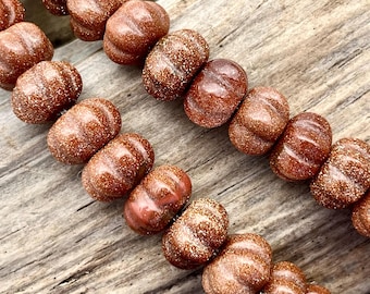 May include: Close-up of a strand of reddish-brown, barrel-shaped beads with a glittery, speckled appearance. The beads are arranged in rows on a weathered wooden surface, showcasing their textured details and warm color.