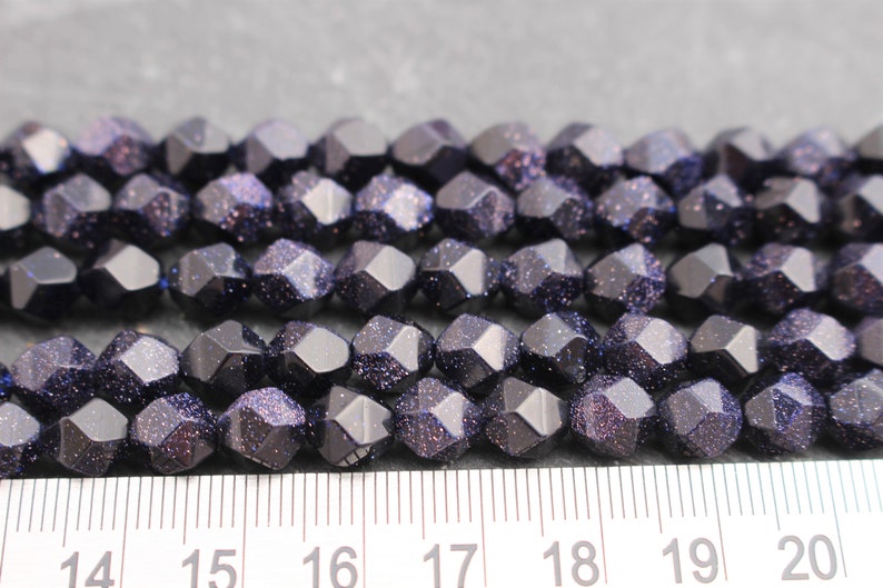 Pode incluir: Close-up de um fio de contas facetadas azul escuro com inclus&otilde;es brilhantes. As contas est&atilde;o dispostas em fileiras ordenadas, e uma r&eacute;gua &eacute; vis&iacute;vel na parte inferior, indicando o seu tamanho. Estas contas s&atilde;o adequadas para fazer joias.