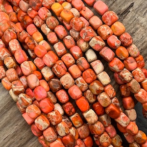 May include: Close-up of strands of orange and pink gemstone beads. The beads are rectangular with rounded edges and have a mottled appearance. The color palette includes shades of orange, pink, and cream, creating a warm and earthy aesthetic. Ideal for jewelry making.