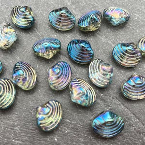 May include: A collection of iridescent, shell-shaped beads. The beads have a textured, ridged surface and display a range of colors including blue, green, and gold. They are scattered on a dark gray surface.