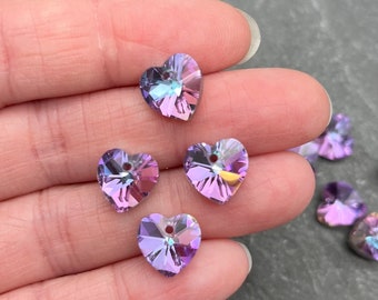 May include: Close-up of several iridescent heart-shaped crystal beads. The beads are a mix of purple, pink, and blue hues, with a hole at the top for stringing. The crystals reflect light, creating a sparkling effect. These beads are ideal for jewelry making.