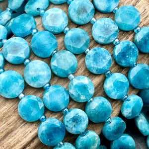 May include: Close-up of a necklace featuring faceted, round, turquoise beads. The beads are strung together with small, clear beads. The necklace is displayed on a wooden surface, showcasing the vibrant blue color and texture of the gemstones.