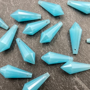 May include: A collection of light blue, faceted crystal pendants. Each pendant has a pointed end and a small hole for stringing. The pendants are a pale, translucent blue and are scattered across a dark surface.