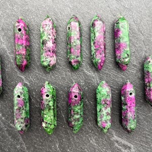 May include: A collection of eleven polished gemstone pendants. Each pendant is a mix of green and magenta colors, with a hole drilled through the top. The pendants are arranged on a gray surface.