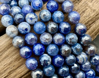 Pode incluir: Close-up de uma coleção de contas de pedras preciosas azuis facetadas. As contas variam em tons de azul, de claro a escuro, com uma aparência texturizada e martelada. As contas são redondas e têm um acabamento polido.
