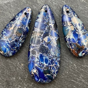 May include: Three teardrop-shaped blue jasper pendants with a hole drilled at the top. The stones have a mosaic pattern of dark and light blue, white, and brown. These pendants are suitable for jewelry making.