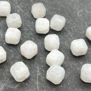 May include: A collection of faceted, white gemstone beads. The beads have a slightly translucent appearance and a geometric shape. They are scattered on a dark, textured surface, showcasing their individual forms and subtle variations.