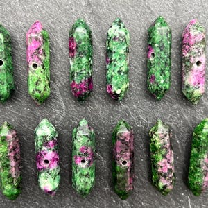 May include: A collection of green and pink gemstone pendants. Each pendant is a cylindrical shape with a hole drilled through the center. The gemstones have a speckled appearance, with a mix of green and pink colors.
