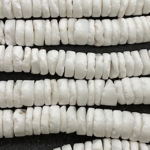 May include: Close-up of multiple strands of white, disc-shaped beads. The beads are strung together, creating a textured, layered effect. The beads appear to be made of a smooth, opaque material. The background is a dark, neutral color.