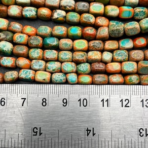 Pu&ograve; includere: Primo piano di una fila di perline quadrate nei toni del turchese, arancione, marrone e verde. Un righello di metallo &egrave; posto sotto, mostrando le misure in centimetri. Le perline hanno un aspetto naturale e terroso.