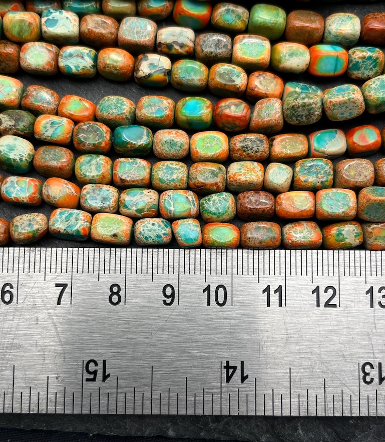 Pu&ograve; includere: Primo piano di una fila di perline quadrate nei toni del turchese, arancione, marrone e verde. Un righello di metallo &egrave; posto sotto, mostrando le misure in centimetri. Le perline hanno un aspetto naturale e terroso.