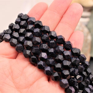 Pode incluir: Um fio de contas facetadas de arenito azul escuro com inclus&otilde;es brilhantes. As contas s&atilde;o seguradas em uma m&atilde;o, mostrando sua forma geom&eacute;trica e cor. As contas t&ecirc;m aproximadamente 1,3 cm de di&acirc;metro, enfiadas juntas.