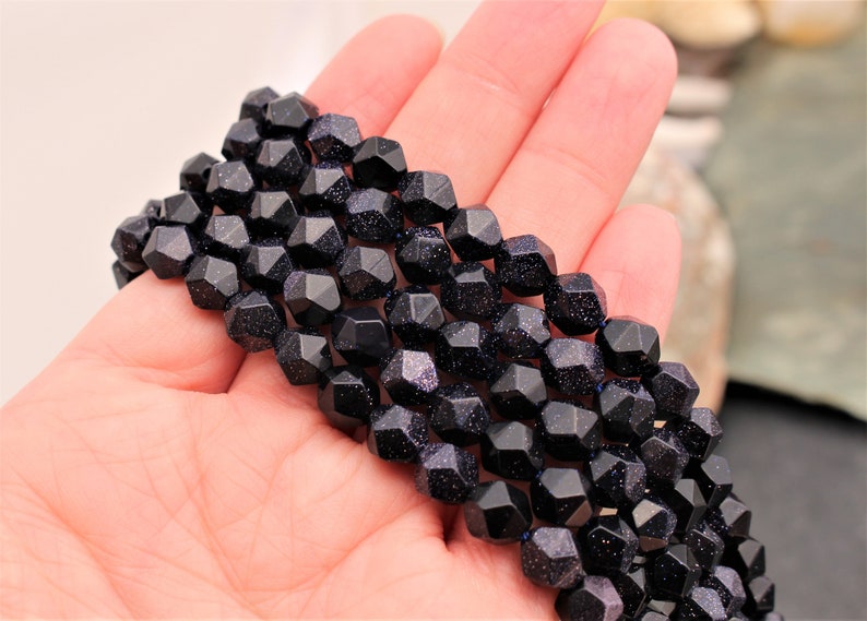 Pode incluir: Um fio de contas facetadas de arenito azul escuro com inclus&otilde;es brilhantes. As contas s&atilde;o seguradas em uma m&atilde;o, mostrando sua forma geom&eacute;trica e cor. As contas t&ecirc;m aproximadamente 1,3 cm de di&acirc;metro, enfiadas juntas.