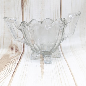 以下が含まれることがあります： 2つのハンドルと3つの脚が付いた透明なガラスの砂糖入れ。ボウルはスカラップの縁とガラスにエッチングされた花の模様があります。砂糖入れは明るい色の木製の表面に置かれています。