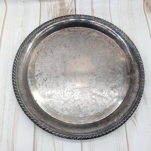 May include: A circular, silver-coloured serving tray featuring an embossed floral pattern in the centre. The tray has a decorative, rope-style edge. The surface shows signs of wear, enhancing its vintage charm. Suitable for serving food or as a decorative item.
