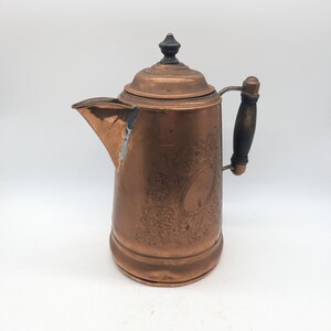 May include: A copper coffee pot with an engraved design and a wooden handle. The pot has a black knob on the lid.