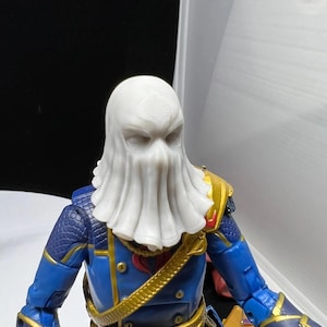 May include: A detailed action figure of a character in a blue and gold uniform with a white hood. The figure has intricate details, including a cobra emblem on the hood and gold accents on the uniform.