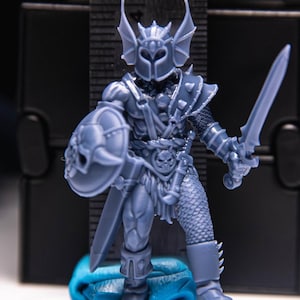 May include: A gray miniature figurine of a warrior in armor, holding a sword and shield. The warrior has a helmet with wing-like features and is standing on a blue base. The figure is highly detailed.