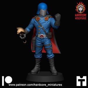 May include: A blue miniature figurine of a character wearing a helmet, a long coat with gold buttons, a red-lined cape, and black boots. The figure holds a gold-colored object in one hand and features a silver belt buckle with a red emblem.