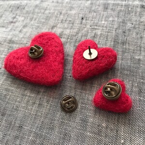 May include: Three red felt heart-shaped pins with metal clasps on the back. The hearts are different sizes.