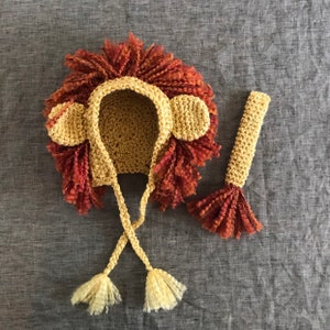 May include: A crocheted lion costume for a baby or toddler. The costume includes a hat with a mane, ears, and a tail. The hat is yellow and the mane is red and brown. The tail is yellow with a red and brown tip.