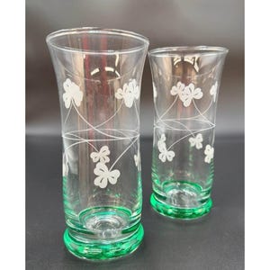 May include: Two clear glass vases with a green base and rim. The vases are decorated with white clover designs and curved lines. The vases are empty and sit on a dark surface.