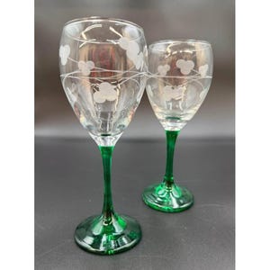 May include: Two clear wine glasses with green stems and bases. The glass bowls are decorated with a white floral design. The glasses are displayed on a dark surface.