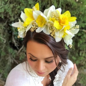 May include: A floral crown featuring yellow and white daffodils and small white flowers. The crown is set against a backdrop of green foliage. The flowers are arranged in a circular pattern, creating a natural and elegant headpiece.