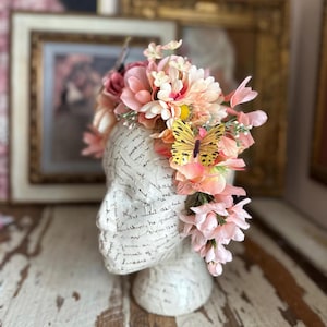 May include: A decorative head bust adorned with a floral crown. The crown features pink and peach-colored flowers, a yellow butterfly, and greenery. The bust is white with script writing.